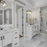 Remodeled primary bathroom with white dual vanities, marble tile flooring, and a glass walk-in shower showing finished bathroom renovation results.