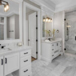 Remodeled primary bathroom with white dual vanities, marble tile flooring, and a glass walk-in shower showing finished bathroom renovation results.